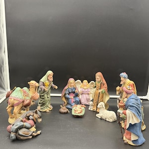 May include: A nativity scene set featuring hand-painted figurines including a central infant in a manger, a kneeling figure in blue, an angel, a shepherd, three wise figures, a camel with a green saddle, and a donkey lying on the ground.