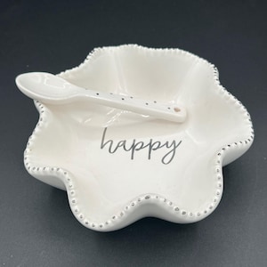 Puede incluir: Un plato de cerámica blanco con borde festoneado y una cuchara pequeña. El plato tiene la palabra "happy" en escritura gris y está decorado con pequeños puntos plateados alrededor del borde y en la cuchara.