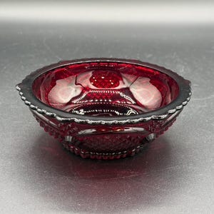 Puede incluir: Un cuenco de cristal rojo rubí profundo con un patrón de diamantes texturizado en el exterior y un borde festoneado. El cristal translúcido refleja la luz, resaltando las complejas facetas geométricas y la forma circular del pequeño plato decorativo.