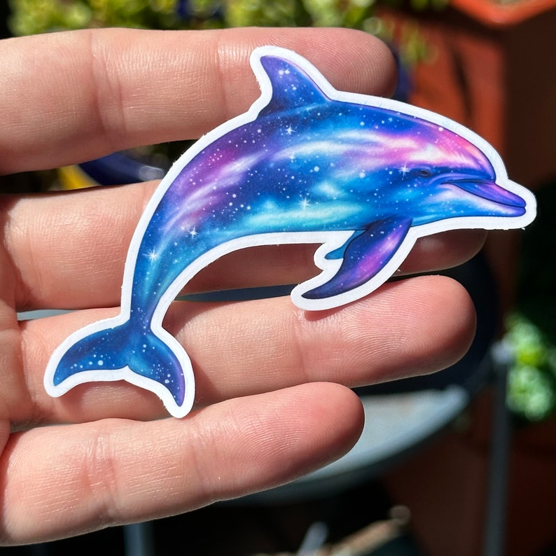 May include: A blue and purple dolphin sticker with a galaxy design. The dolphin is jumping out of the water with its tail in the air. The galaxy design is made up of stars and swirls of color.