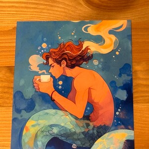 Może przedstawiać: Aquarela przedstawiająca syrenę z brązowymi włosami i ogonem w zielone, niebieskie i ż&oacute;łte łuski. Syrena trzyma biały kubek, z kt&oacute;rego unosi się para.