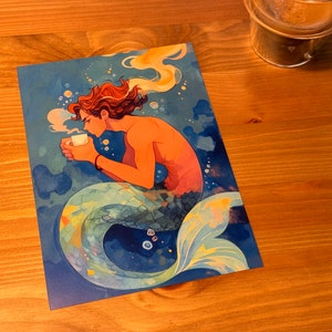 Może przedstawiać: Aquarela przedstawiająca syrenę z niebieskimi, zielonymi i ż&oacute;łtymi łuskami, trzymającą białą filiżankę kawy. Syrena ma brązowe włosy i siedzi w niebiesko-białym otoczeniu oceanu.