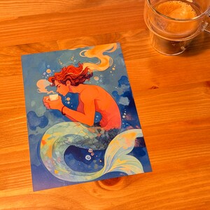 Może przedstawiać: Aquarelowe ilustracje syreny z pomarańczowymi włosami trzymającej białą filiżankę kawy. Syrena siedzi na niebiesko-zielonym ogonie z ż&oacute;łtymi i pomarańczowymi akcentami. Tło to niebiesko-zielony akwarelowy wash z białymi bąbelkami.