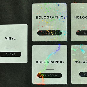 May include: Four vinyl sticker samples with different holographic finishes. The top left sticker says "VINYL" and "CLEAR". The top right sticker says "HOLOGRAPHIC" and "GLITTER". The bottom left sticker says "HOLOGRAPHIC" and "RAINBOW". The bottom right sticker says "HOLOGRAPHIC" and "GEMS".