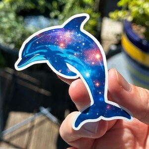 May include: A blue dolphin sticker with a galaxy pattern. The dolphin is jumping out of the water and has a white outline.