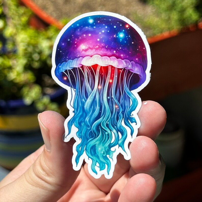 May include: A colorful sticker of a jellyfish with a galaxy design. The jellyfish is blue, purple, and pink with a swirling galaxy pattern in the bell. The tentacles are long and flowing.