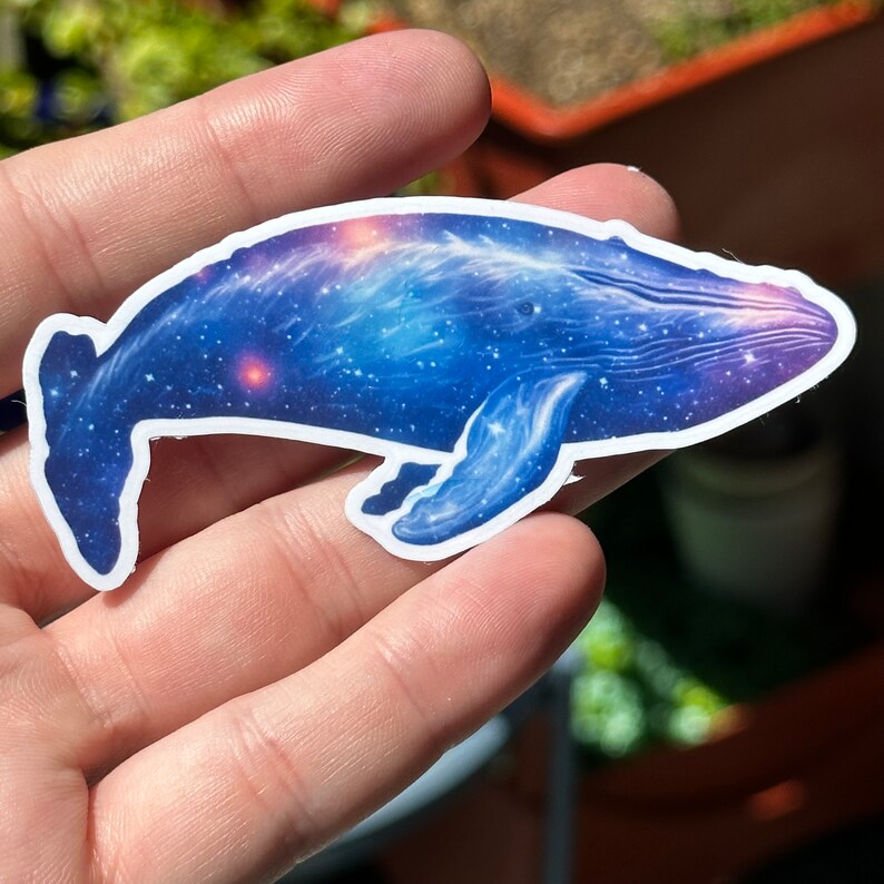 May include: A blue whale sticker with a galaxy design. The whale is swimming with its mouth open and has a white outline.