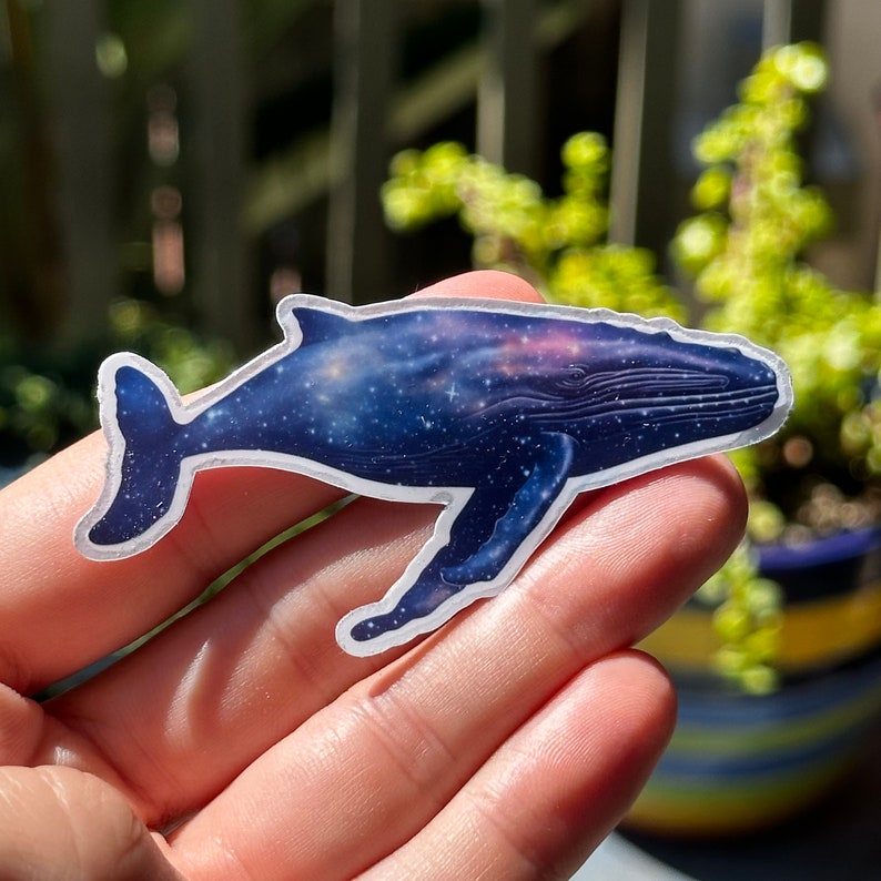 May include: A whale sticker with a galaxy design. The whale is blue and white with a dark blue galaxy pattern. The sticker is on a hand.