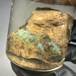 May include: A rough, brown and green rock with iridescent blue and green opal inclusions is displayed in a clear glass jar with a wooden lid.