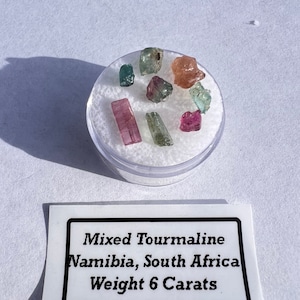 May include: A small container of mixed tourmaline gemstones from Namibia, South Africa. The label reads "Mixed Tourmaline Namibia, South Africa Weight 6 Carats".