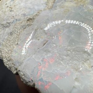 May include: A rough, white and grey rock with a red and iridescent opal inside. The opal has a rainbow sheen.