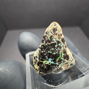 May include: A rough, triangular-shaped gemstone with a dark, textured surface. The stone displays vibrant flashes of green and blue, set against a black matrix. It rests on a clear, rectangular display stand, showcasing its natural beauty.