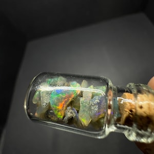 May include: A small glass bottle filled with iridescent opal chips. The bottle has a cork stopper and is filled with a variety of opal pieces.