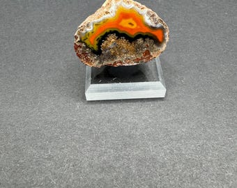 May include: A polished agate geode with a natural, irregular shape. The geode is cut in half, revealing a colorful interior with bands of orange, black, and white. The geode is displayed on a clear acrylic stand.