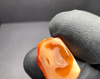 May include: A close-up of a small, orange and white, irregularly shaped, polished stone. The stone has a smooth, glossy surface and is held in a gloved hand.