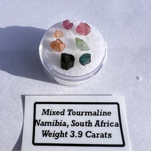 May include: A collection of mixed tourmaline gemstones in a clear plastic container. The gemstones are various colors, including pink, green, and black. A label below reads "Mixed Tourmaline Namibia, South Africa Weight 3.9 Carats."