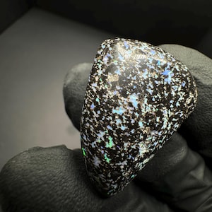 May include: A black opal gemstone with a speckled pattern of iridescent blue, green, and white. The opal is irregularly shaped and has a smooth, polished surface.