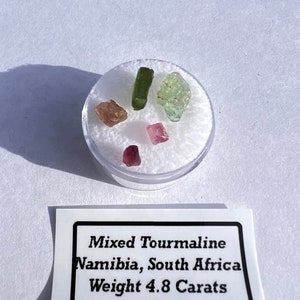 May include: A small container of mixed tourmaline gemstones from Namibia, South Africa. The gemstones are pink, green, and light green. The label reads "Mixed Tourmaline Namibia, South Africa Weight 4.8 Carats".