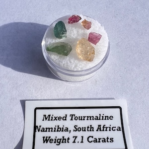 May include: A small container of mixed tourmaline gemstones from Namibia, South Africa. The gemstones are green, pink, and yellow. The label reads "Mixed Tourmaline Namibia, South Africa Weight 7.1 Carats".