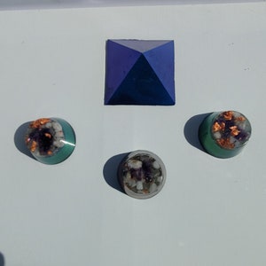 May include: Three small resin pyramids with amethyst and copper flakes embedded inside. The pyramids are on a white surface. The pyramid in the center is a darker shade of blue than the two on the sides.