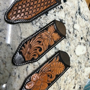 May include: Three leather knife sheaths with different designs. The sheaths are black with brown leather accents. The designs include a woven pattern, a floral pattern, and a leaf pattern.