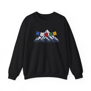 May include: A black crewneck sweatshirt featuring a graphic print of a snow-capped mountain peak. A string of five colorful prayer flags in blue, white, red, green, and yellow is draped across the front of the mountain illustration.