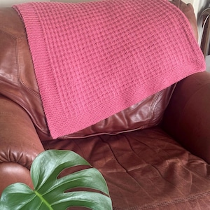 May include: A solid pink knitted blanket featuring a textured waffle stitch pattern throughout the center and a smooth, flat-knit border along the edges. The fabric has a consistent, uniform color and a structured, repeating geometric grid design.