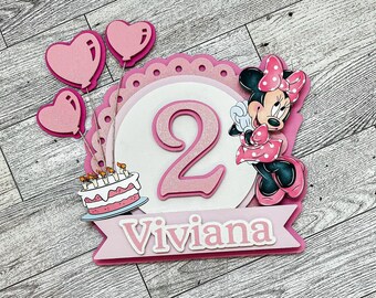 Könnte beinhalten: Rosa-weißer Tortendekor mit einer Zeichentrickfigur von Minnie Mouse, einer rosa Zahl 2, einer rosa Torte mit Kerzen und drei rosa Herzluftballons. Der Dekor enthält auch den Namen "Viviana" in rosa Buchstaben.