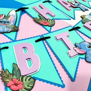 Puede incluir: Una colorida pancarta con banderines celeste, rosa y morado. Cada bander&iacute;n tiene un borde festoneado y presenta una letra, con im&aacute;genes de Stitch y Lilo, tablas de surf y flores tropicales. Cinta negra conecta los banderines.