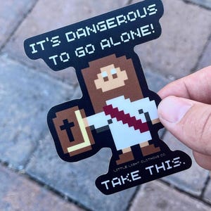 May include: A pixelated sticker featuring a character holding a cross and a book. The sticker has the text "IT'S DANGEROUS TO GO ALONE! TAKE THIS." The character is wearing a white robe with a red sash. The sticker is black with white text.