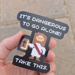 May include: A black sticker with pixelated art of a figure holding a book and wearing a white robe. The sticker reads "IT'S DANGEROUS TO GO ALONE! TAKE THIS."