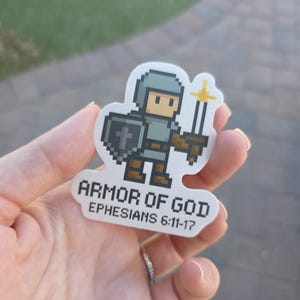 May include: A sticker featuring a pixelated knight in armor, holding a sword and shield with a cross. The text "ARMOR OF GOD" and "EPHESIANS 6:11-17" are printed below the knight. The sticker is white with a die-cut shape.