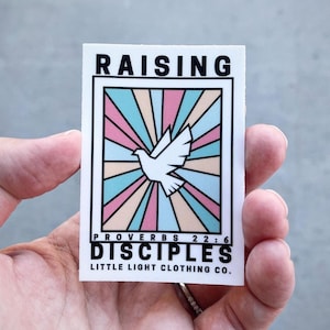 May include: A white sticker with a colorful design featuring a dove and the text "Raising Disciples" with the scripture reference "Proverbs 22:6" and the text "Little Light Clothing Co."