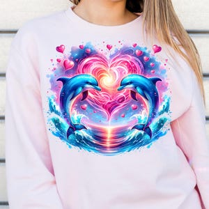 Puede incluir: Sudadera rosa claro con un gr&aacute;fico vibrante de dos delfines azules saltando de las olas, formando un coraz&oacute;n. Corazones rosas y un fondo de puesta de sol colorido completan el dise&ntilde;o.
