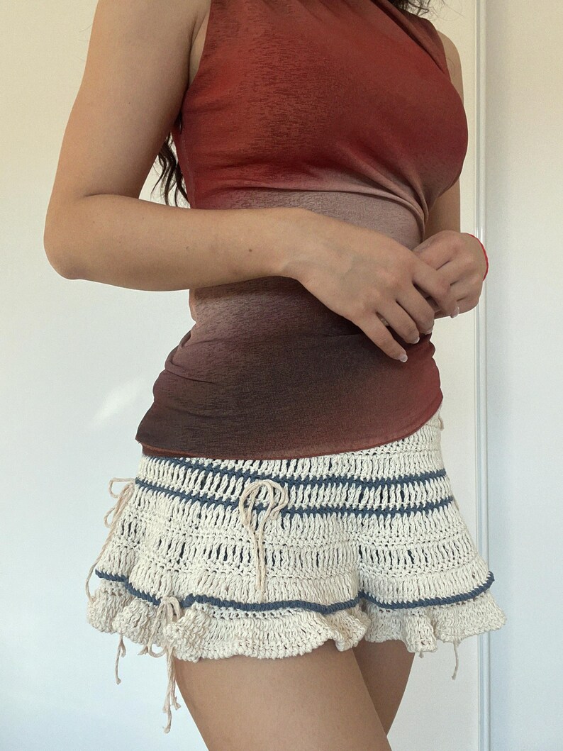 May include: A sleeveless top with a gradient of red to brown tones. Paired with a cream-colored crochet skirt with blue stripes and a ruffled hem. The skirt has drawstrings at the waist.