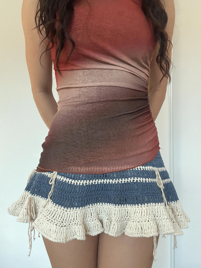 May include: A two-piece outfit featuring a sleeveless, ombre-dyed top in shades of red and brown. The skirt is a blue and cream crochet mini skirt with a ruffled hem and drawstring details. The outfit is a stylish summer ensemble.