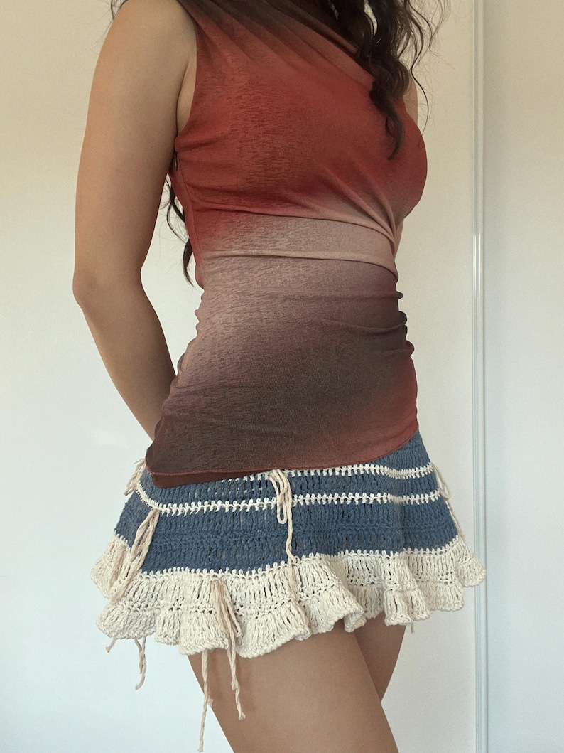 May include: A sleeveless, form-fitting top with a gradient of red to brown hues. Paired with a blue and cream crochet mini skirt with a ruffled hem and drawstring details. A stylish, unique outfit.