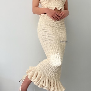 May include: Cream-colored crochet two-piece set. The set includes a strapless top and a fitted midi skirt with a ruffled hem. The outfit is paired with cream-colored high heels. The image is taken in front of a plain white wall.