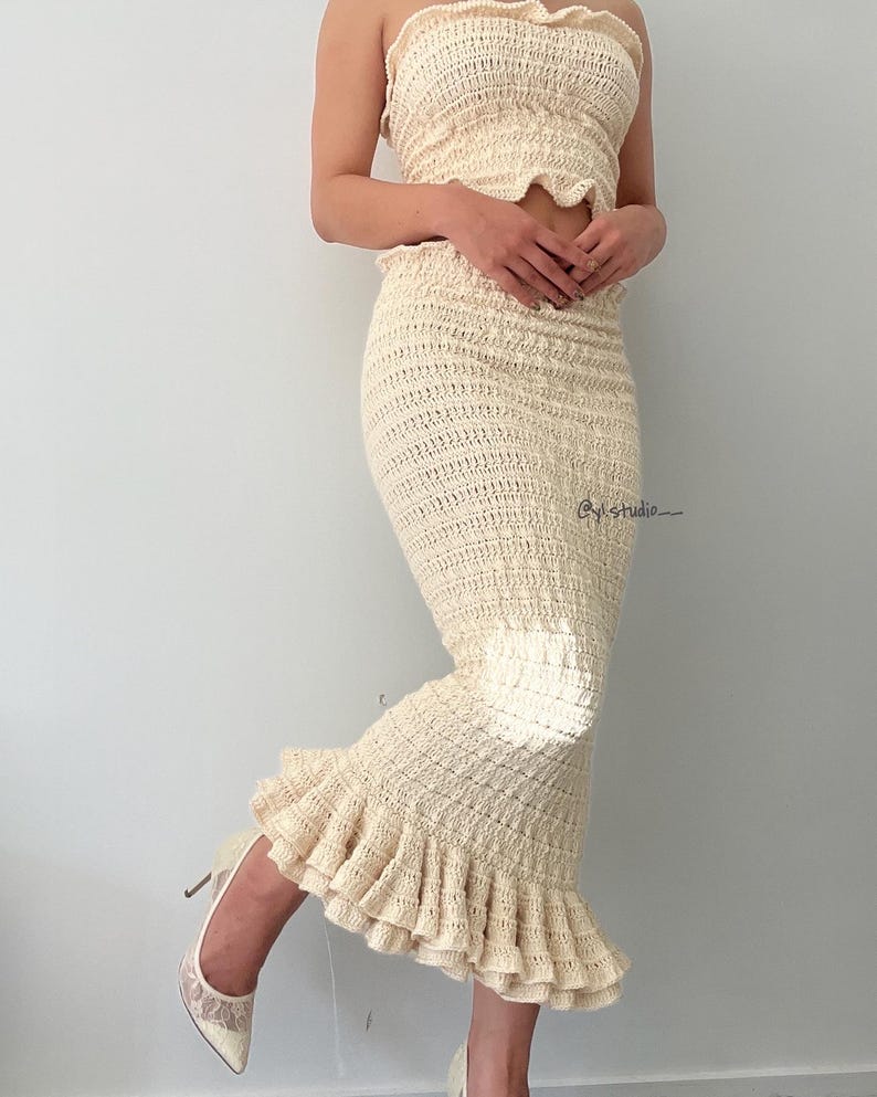 May include: Cream-colored crochet two-piece set. The set includes a strapless top and a fitted midi skirt with a ruffled hem. The outfit is paired with cream-colored high heels. The image is taken in front of a plain white wall.