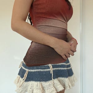 May include: A sleeveless top with a gradient of red to brown tones, paired with a blue and cream crochet mini skirt. The skirt features horizontal blue stripes and a ruffled cream-colored hemline, with cream-colored ties.