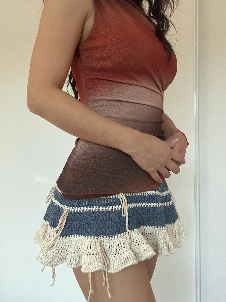 May include: A sleeveless top with a gradient of red to brown tones, paired with a blue and cream crochet mini skirt. The skirt features horizontal blue stripes and a ruffled cream-colored hemline, with cream-colored ties.