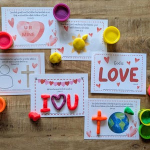 May include: A collection of colorful playdough and cards with religious themes. The cards feature phrases like "God is LOVE" and "UR MINE," along with Bible verses and heart illustrations. Playdough containers in various colors are scattered around.