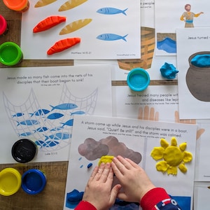 Puede incluir: Un colorido juego de actividades educativas con historias bíblicas. La imagen muestra páginas impresas con ilustraciones y texto, junto con plastilina de varios colores. La escena incluye pan, peces y una red, con las manos de un niño formando una sol.