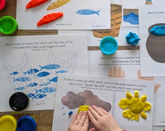 May include: A colorful educational activity set featuring Bible stories. The image shows printed pages with illustrations and text, along with modeling clay in various colors. The scene includes bread, fish, and a net, with a child's hands shaping yellow clay into a sun.