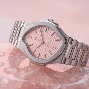 Puede incluir: Un reloj de pulsera de acero inoxidable con esfera rosa. El reloj tiene una forma de caja única y una correa de metal. La esfera presenta un patrón texturizado y el texto "TIME BUILT DIFFERENTLY".