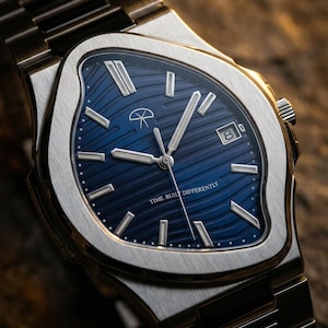 Puede incluir: Reloj de pulsera de acero inoxidable con esfera azul con un patrón de ondas. El reloj tiene agujas e indicadores plateados, una ventana de fecha y el texto "TIME BUILT DIFFERENTLY". La correa del reloj también es de acero inoxidable.