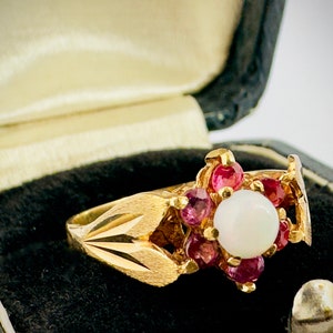 May include: A gold ring featuring a white opal surrounded by pink gemstones. The ring is displayed in a black velvet ring box.