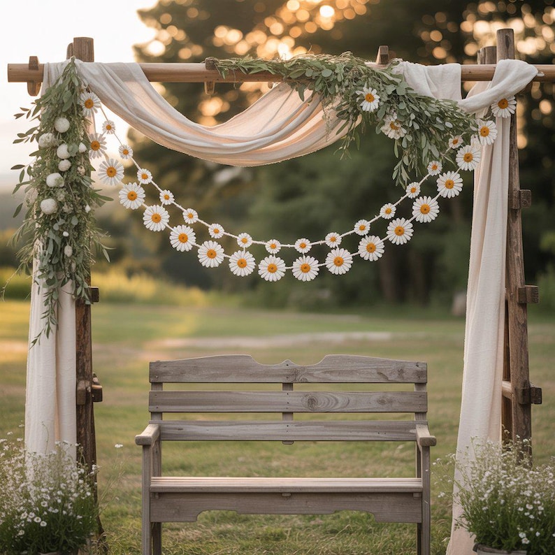 May include: A wooden wedding arch decorated with white fabric, greenery, and a daisy garland. A wooden bench sits in front of the arch, with a backdrop of a grassy field and trees. The scene is bathed in soft, natural light.