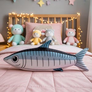 Könnte beinhalten: Ein kissenförmiger Fisch in Blau und Weiß mit schwarzen Details. Es liegt auf einer rosa Bettdecke, im Hintergrund sind Stofftiere zu sehen. Ein dekoratives Element für das Kinderzimmer.