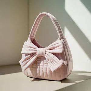 May include: A light pink quilted handbag with a large bow detail. The bag has a rounded shape with vertical stitching and a matching handle. The bow is centered on the front, adding a decorative touch to the accessory.